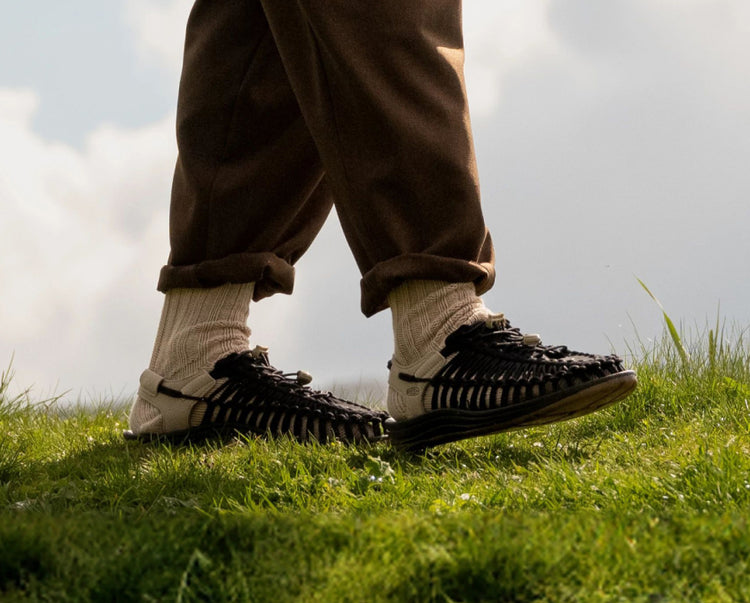 Knee-down short of man wearing tan socks and brown Uneek sandals walking across grass. 