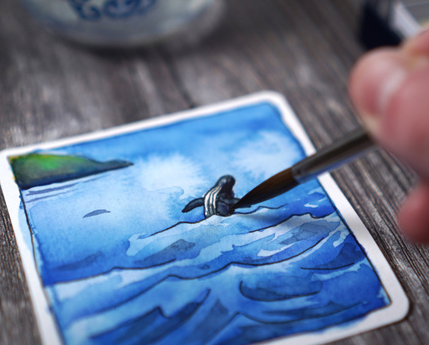 A person painting a small watercolor picture of an ocean scene.