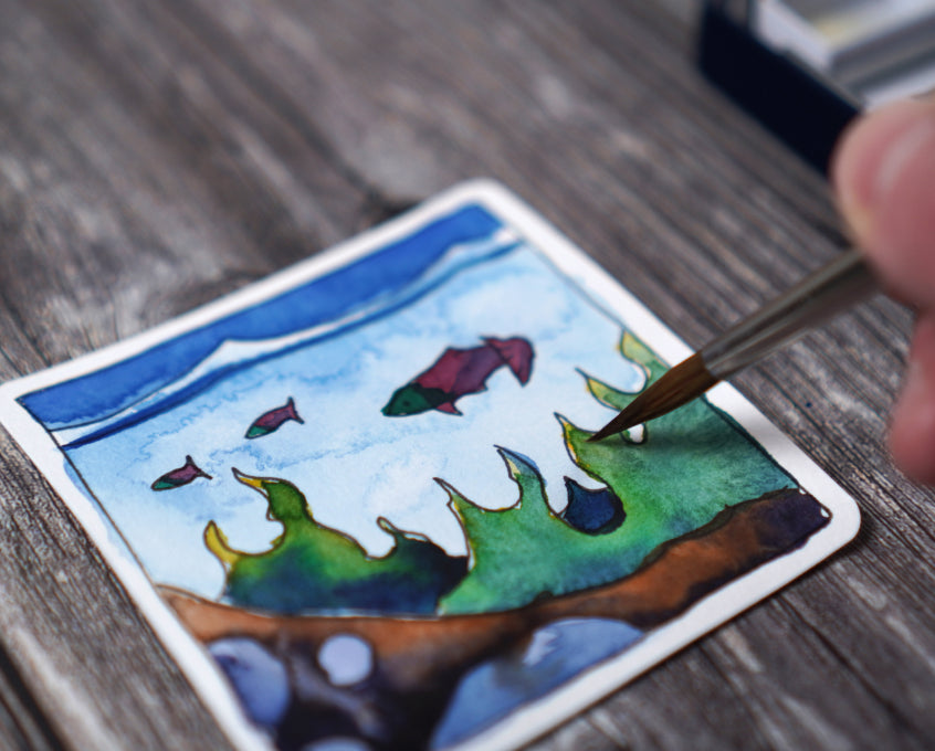 A person painting a small watercolor picture of an underwater scene.