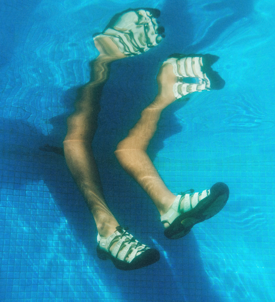 Underwater shot of a person's legs in a blue pool while wearing tan KEEN x Gramicci collaboration Newport sandals