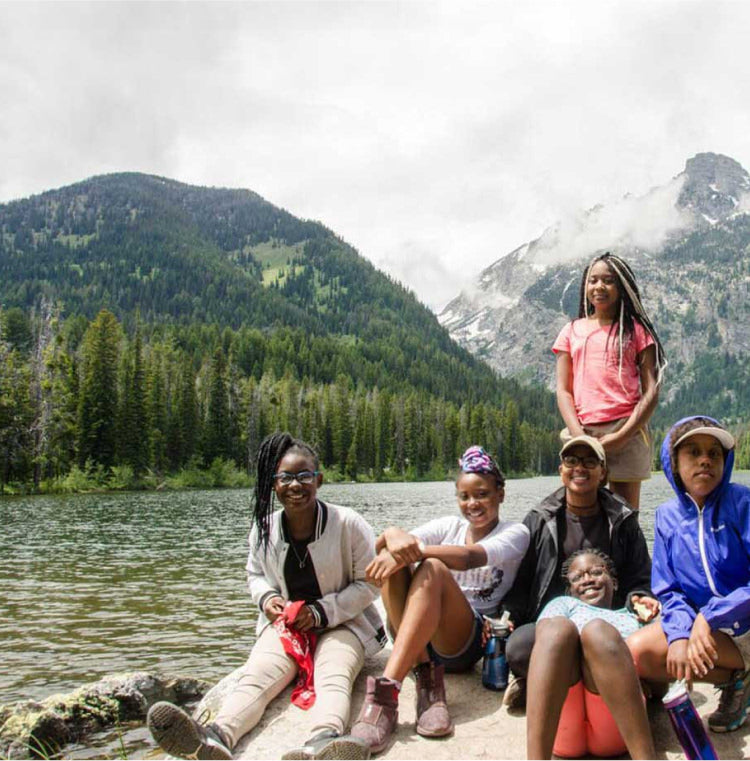group of teens in hiking boots resting near a lake with mountains behind them 