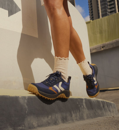 Knee-down shot of woman wearing navy blue KS86 sneakers with tan frilly ankle socks walking in city landscape.
