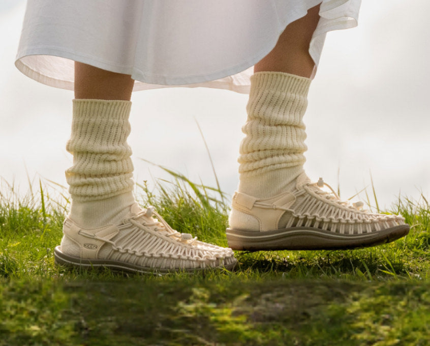 Knee-down shot of woman in white dress and white socks, wearing white Uneek sandals while walking through grass