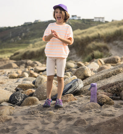 Little kid in purple hat, purple water-bottle and purple Newport Boundless sandals standing on the shore of beach with rocks in the background . 