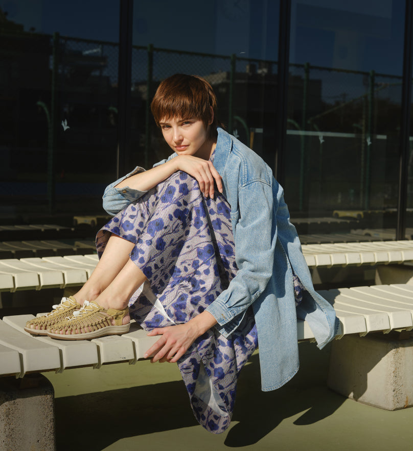 Woman wearing tan Uneek sandals and a blue floral dress sitting with her feet up on a white bench. 