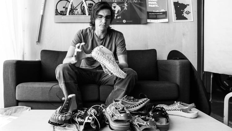 Black and white photo of Man sitting on a couch, holding a mold of the uneek sandal with other uneek prototypes on the table in front of him