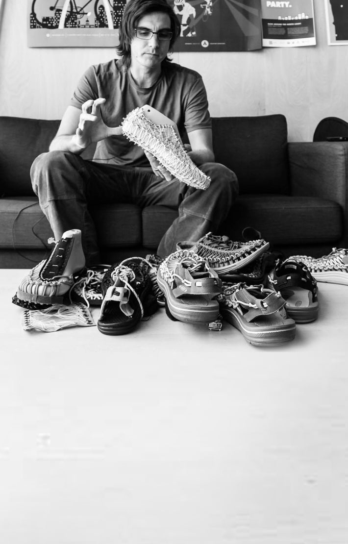 Black and white photo of Man sitting on a couch, holding a mold of the uneek sandal with other uneek prototypes on the table in front of him
