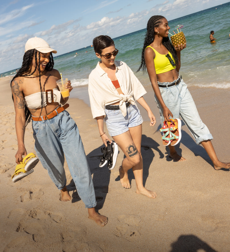 Three women walking along beach shoreline while carrying Elle sandals and drinking juice