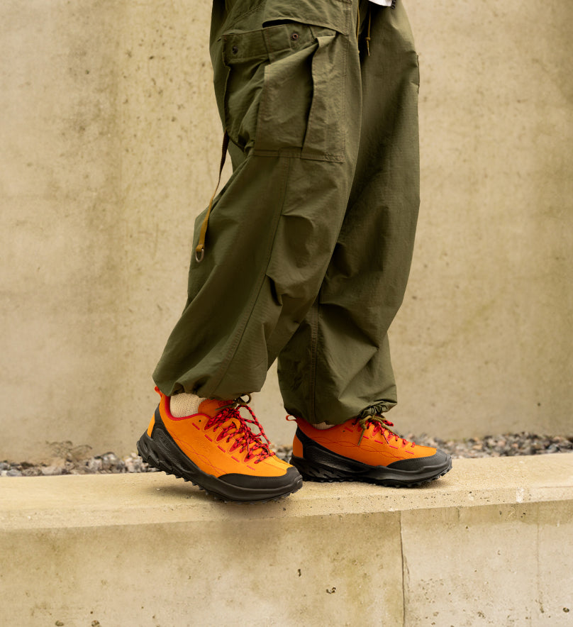 Waist down shot of person in green cargo pants standing on cement wall and wearing orange and black Jasper Zionic sneakers