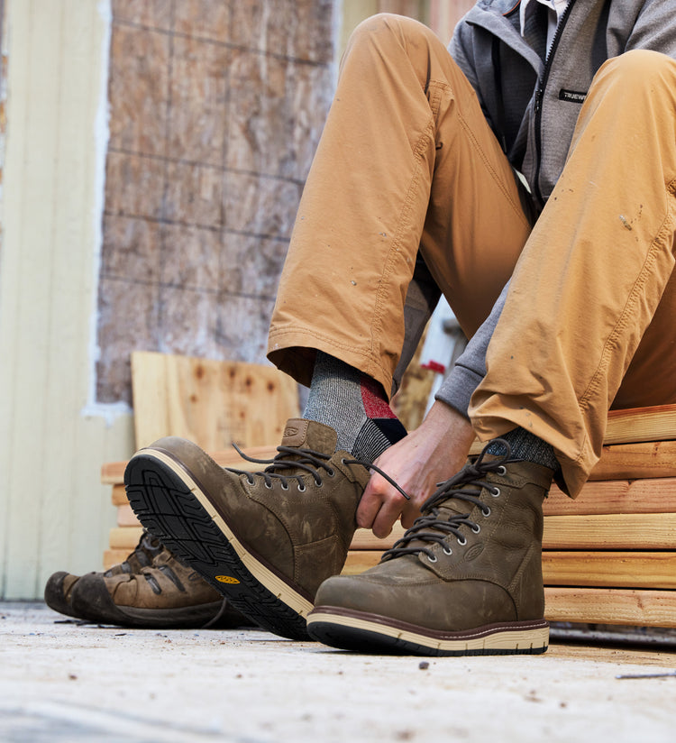 Knee down shot of man sitting on wood planks and lacing up brown San Jose work boots