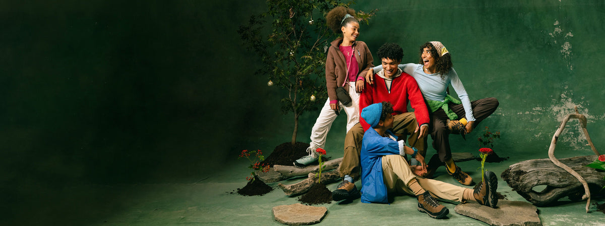 A family of 4 dressed in holiday apparel and posed in front of trees, wood, and rocks while wearing various styles of KEEN sneakers. 