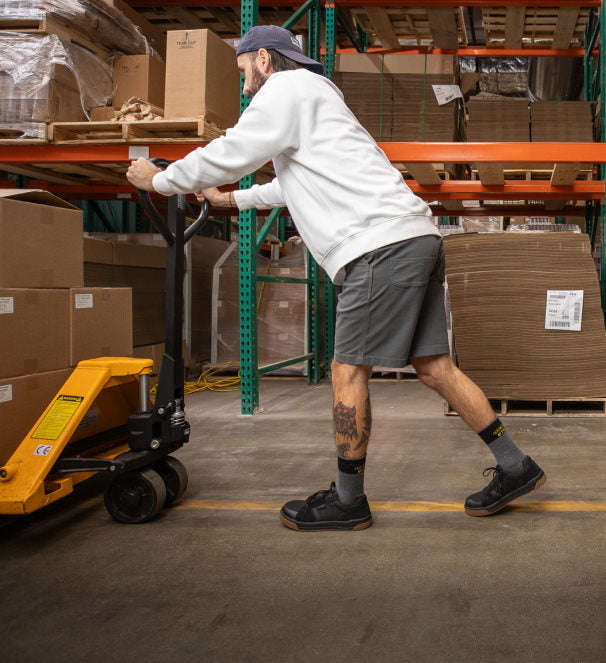 Warehouse worker wearing black Kenton sneakers and pushing a yellow cart full of boxes through warehouse aisles. 