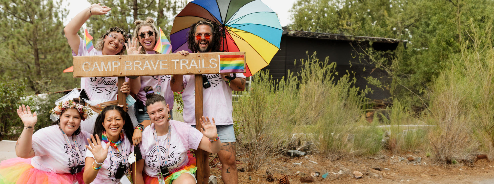  A group of 6 people wearing matching shirts and rainbow gear standing in front of a sign that reads "Camp Brave Trails"