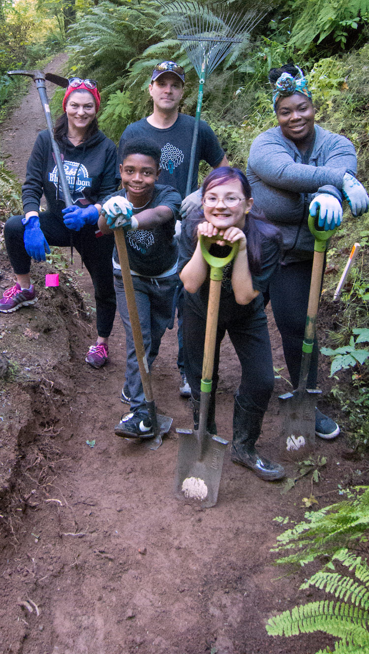 Group of adult and child volunteers holding tools while completing trail work in a forest