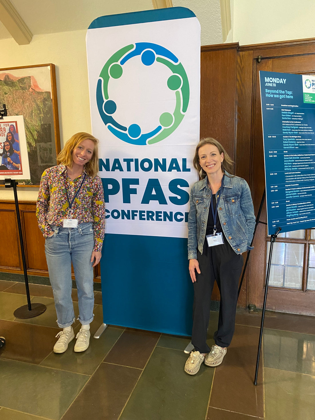 Two women smiling for a photo, standing in front of a sign for the National PFAs Conference.