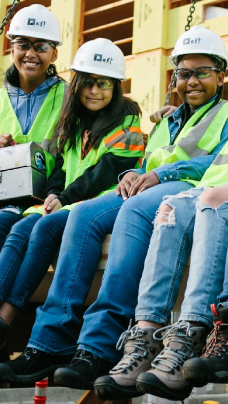 group of five kids wearing jeans, various keen boots, contstructions hats and high visisbility vests sitting on construction site bench