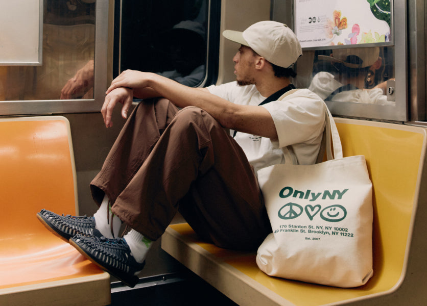 A man sitting in a subway car, wearing KEEN UNEEK sandals and a tote bag.
