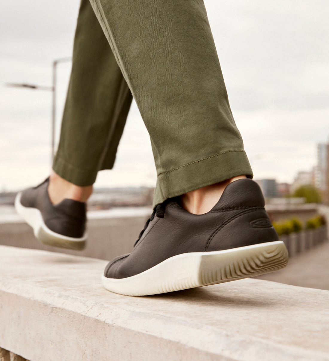 Knee down shot of woman wearing green pants and Black KNX sneakers while walking on cement block 