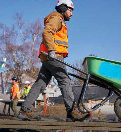 Man in orange visibility vest and protective helmet pushing a wheelbarrow through a construction site while wearing brown and black Evanston work boots.