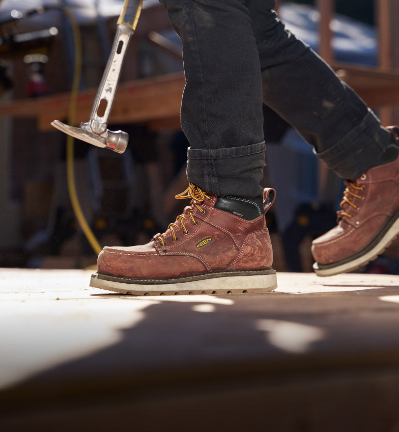 Knee-down shot of man wearing brown Cincinnati work boots and walking across pavement while carrying a hammer. 