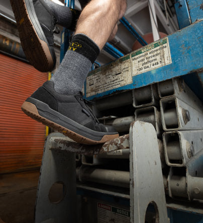 Knee-down shot of man stepping up into forklift while wearing black Kenton low sneakers and gray crew socks. 