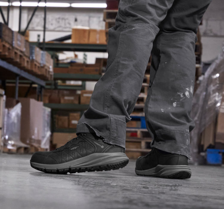 A person wearing a pair of black KEEN work sneakers in a warehouse.
