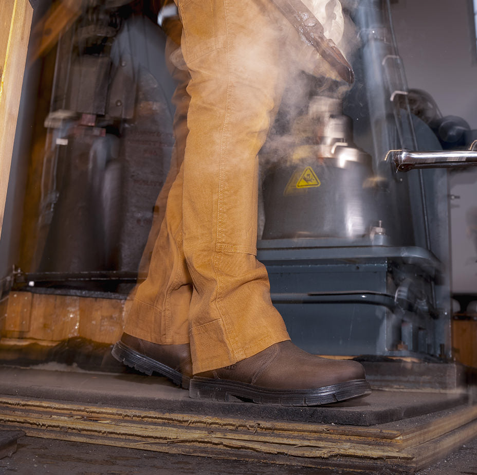 Waist down shot of woman wearing overalls with brown leather San Jose slip on workboots while carrying metal through workshop 