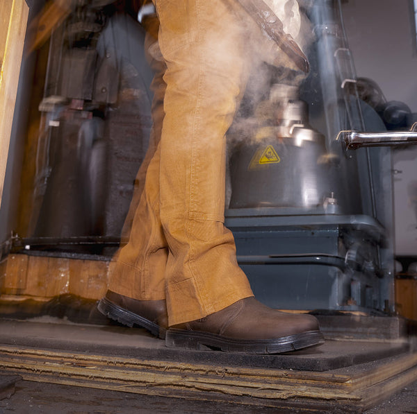 Waist down shot of woman wearing overalls with brown leather San Jose slip on workboots while carrying metal through workshop 