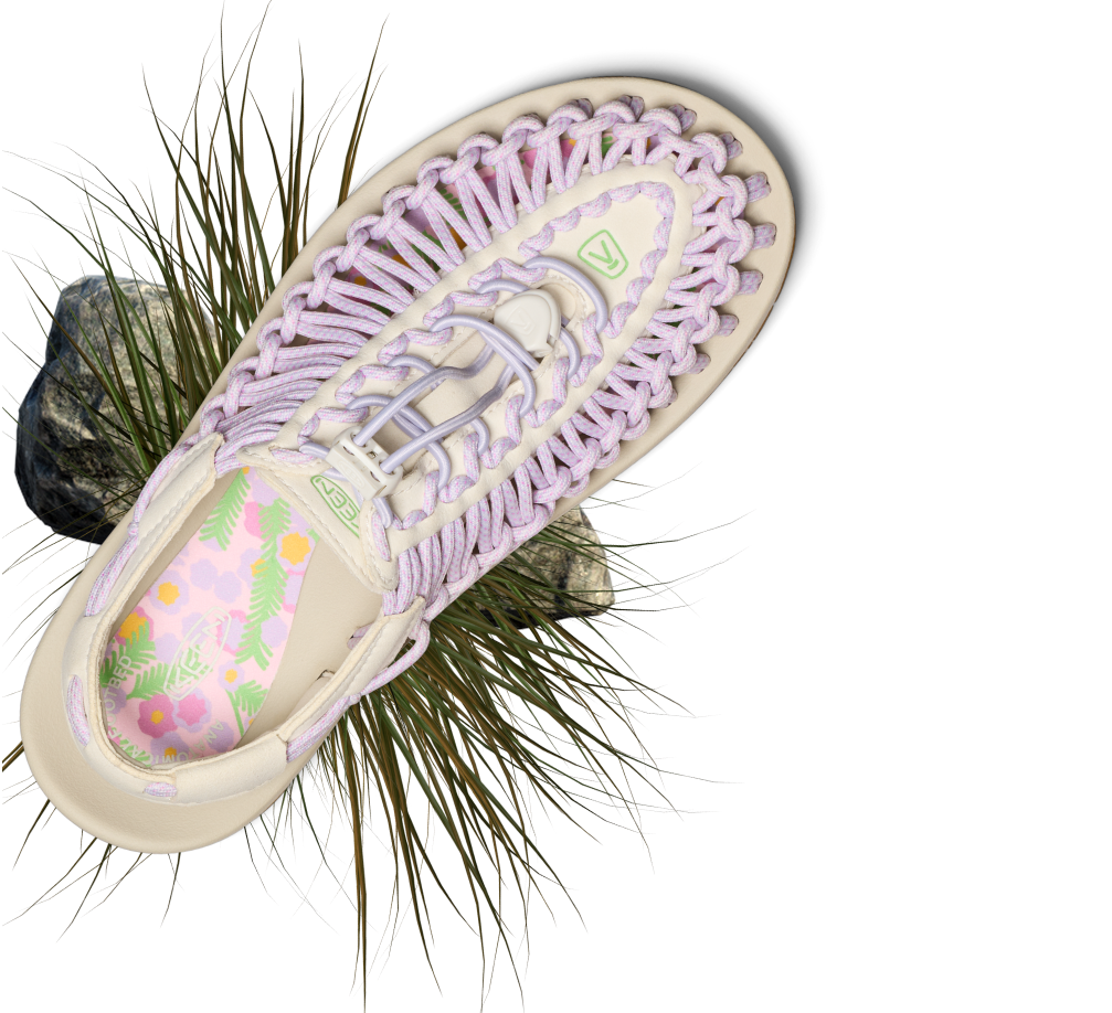 Product shot of lavender and cream uneek open-air sandal with grassy detail surrounding the product. 