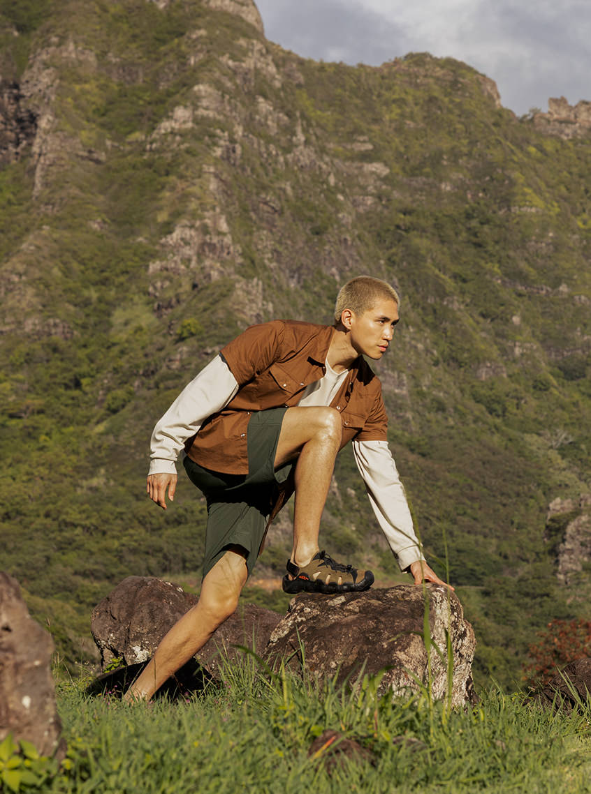 Man in orange shirt and shorts wearing mens leather Hyperport fisherman outdoor sandals and while climbing onto a rock in mountainous setting. 