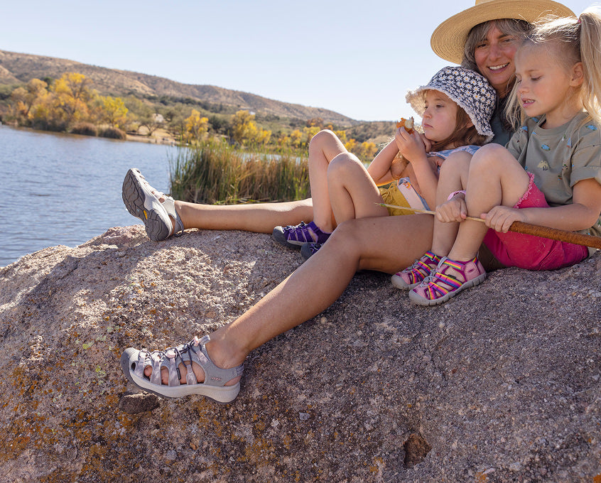 Mom and two young girls all wearing Newport water sandals while sitting on large rock overlooking lake.