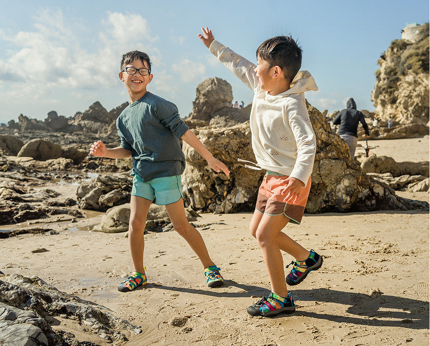 Two young boys wearing Seacamp sandals at beach surrounded by large coastal rocks. 