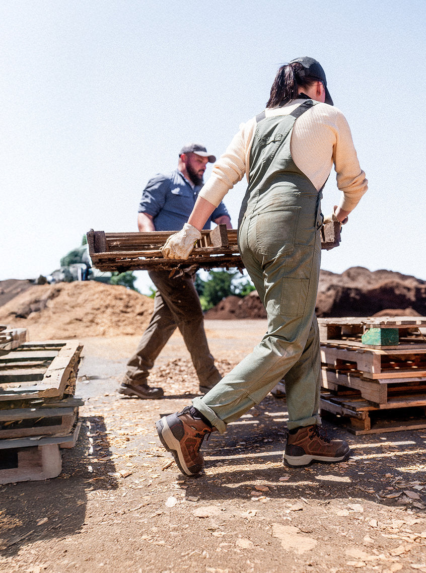 A man and woman in overalls carrying a pallet together while wearing Skokie work boots. 