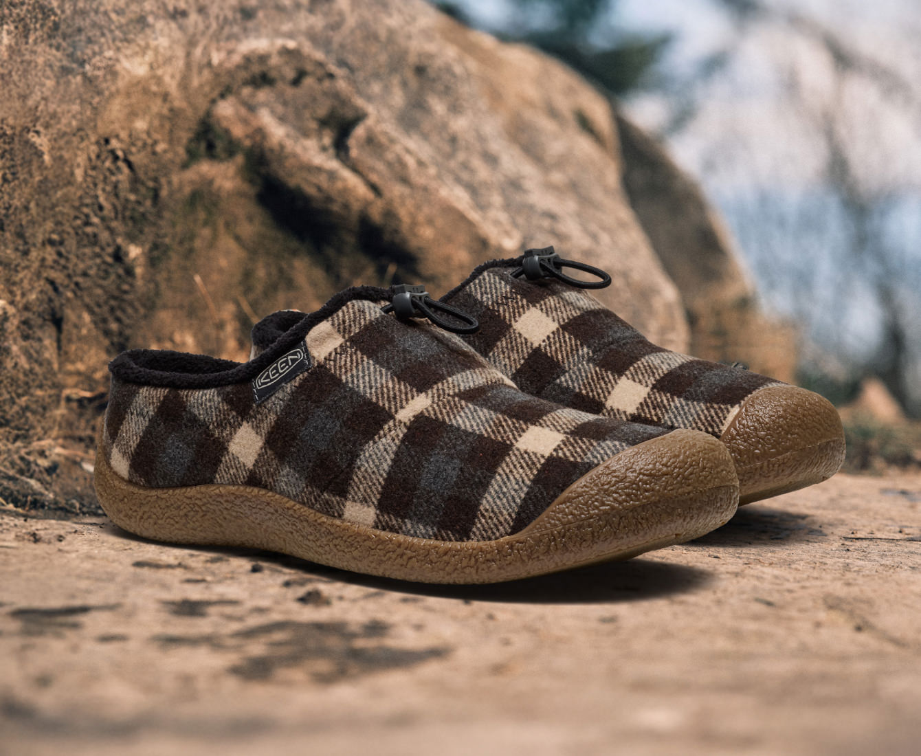 Product shot of brown, tan, cream, and blue plaid Howser slide-in slippers  with outdoor boulder in the background.