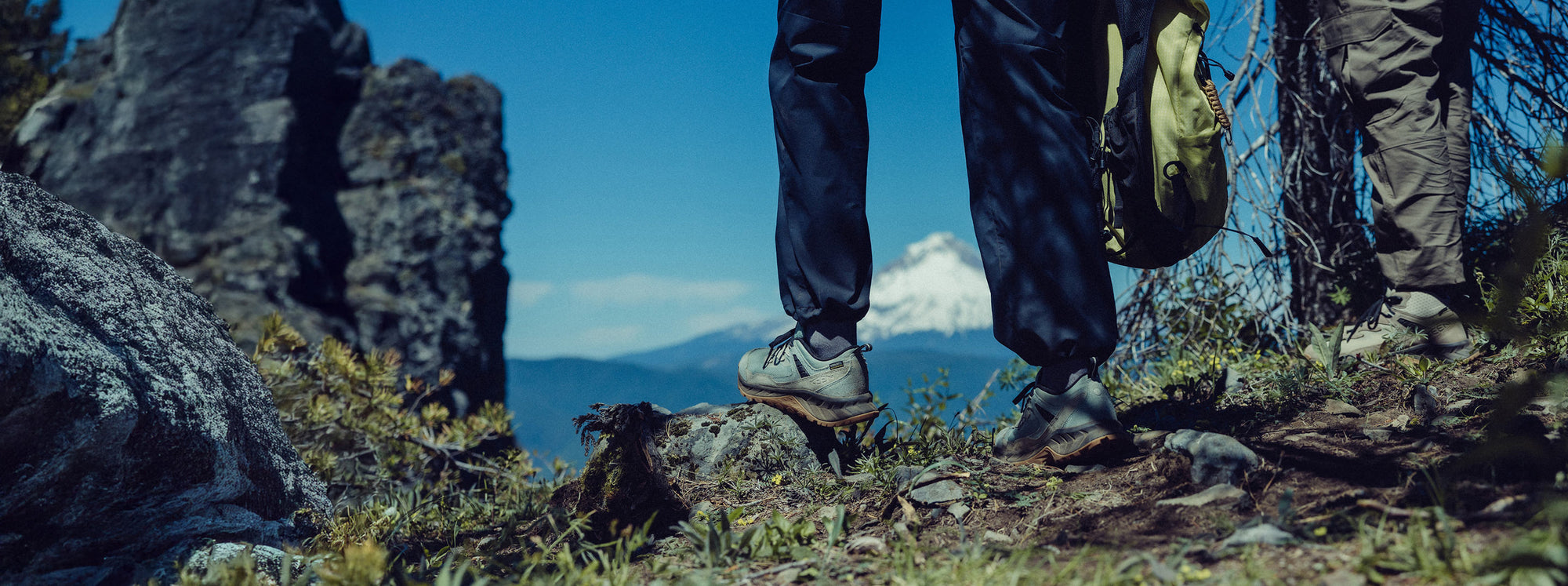 Knee-down shot of Man and woman wearing Hightrail hiking boot and Hightrail hiking shoes while carrying green backpack and looking at a mountain view. 
