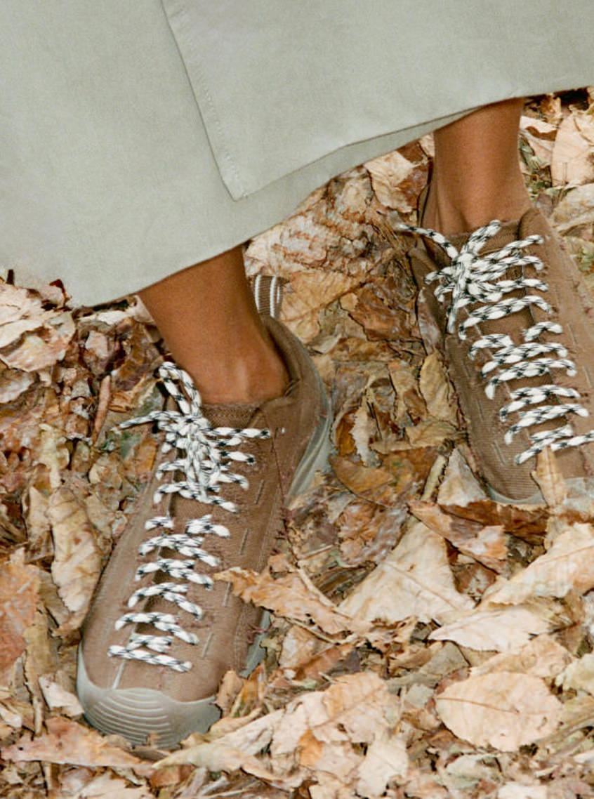 Knee-down shot of woman in long green skirt wearing KEEN and Skall collaboration Jasper sneakers with brown leather and white and green laces on crunchy fall leaves. 