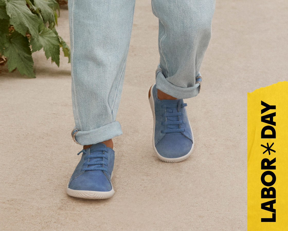 Knee-down shot of kid wearing blue jeans and blue leather sneakers walking across tan concrete. Embedded text reads "Labor Day" in yellow. 