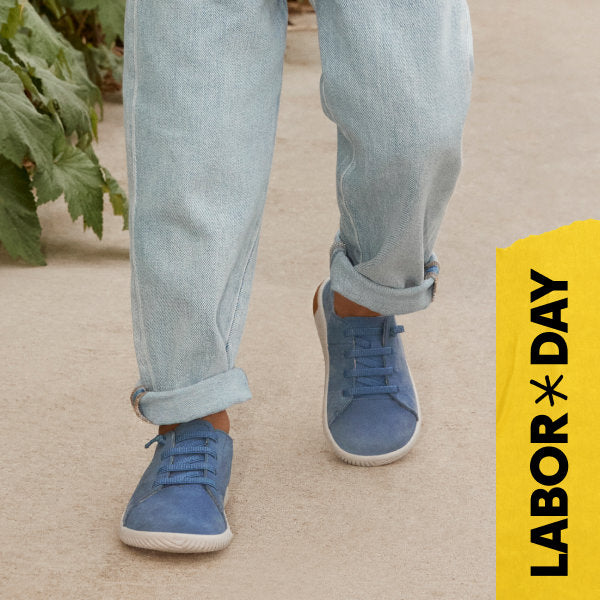 Knee-down shot of kid wearing blue jeans and blue leather sneakers walking across tan concrete. Embedded text reads "Labor Day" in yellow. 
