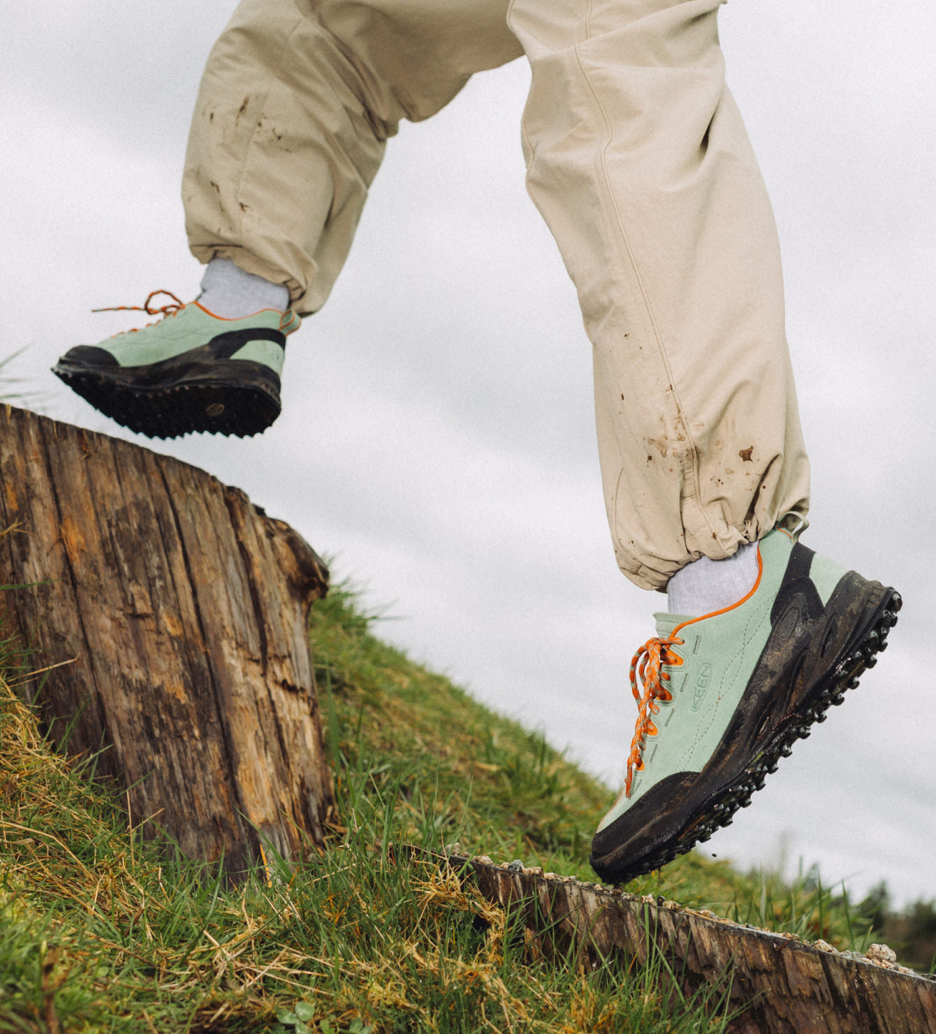Waist down shot of man in turquoise and orange Jasper Zionic sneakers leaping across tree stumps on grassy hillside.