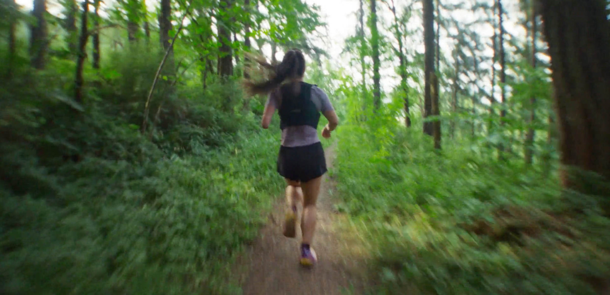 Shot from behind as woman in running vest and long ponytail runs through a dense forest trail.