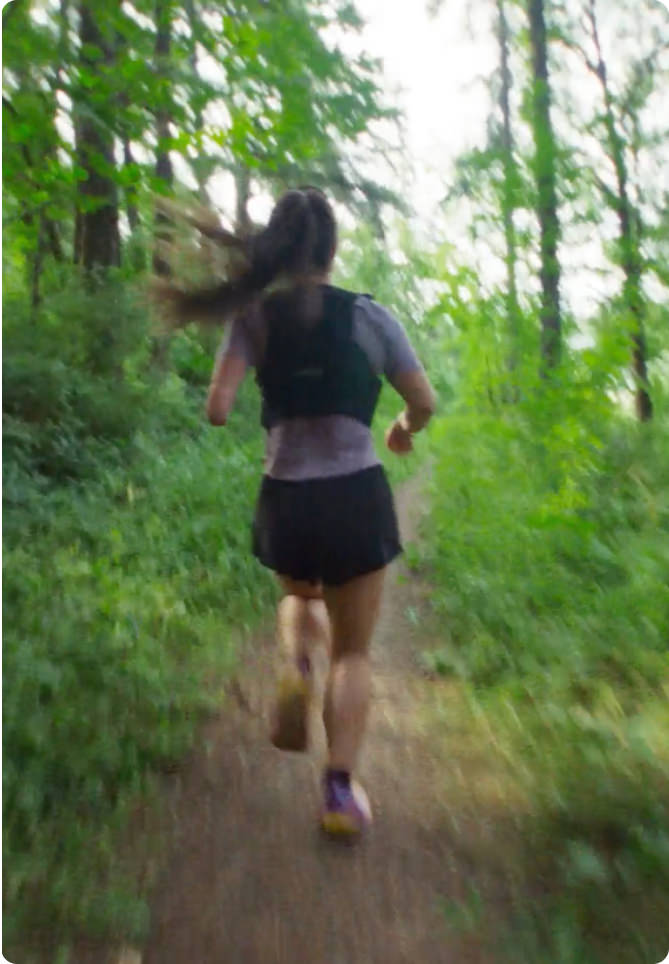 Shot from behind as woman in running vest and long ponytail runs through a dense forest trail.