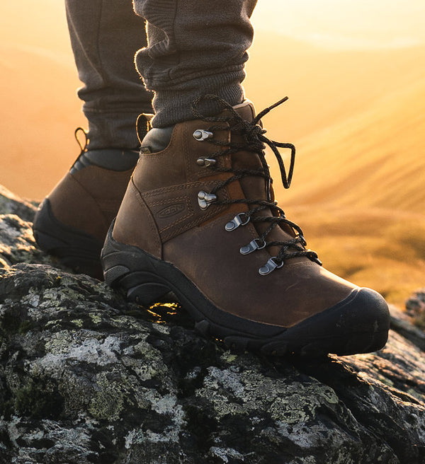 A person wearing a pair of brown and black KEEN hiking boots, standing at the top of a mountain.