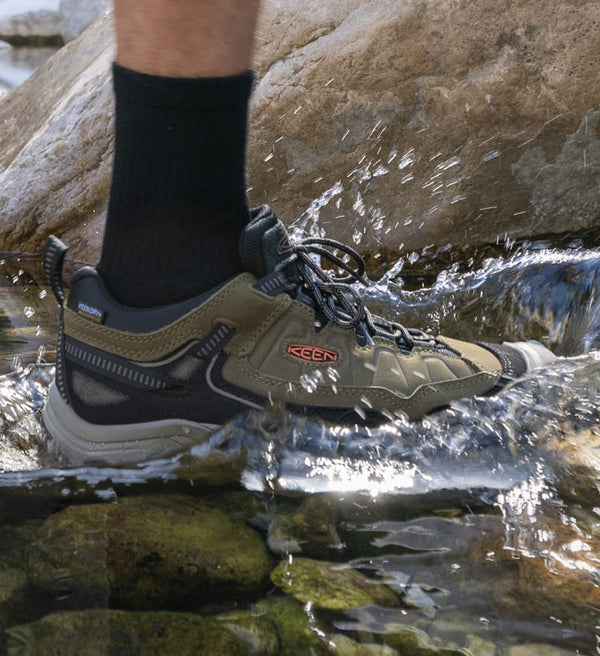Knee-down shot of man wearing waterproof hiking shoes and black crew socks, hiking through rocky stream. 