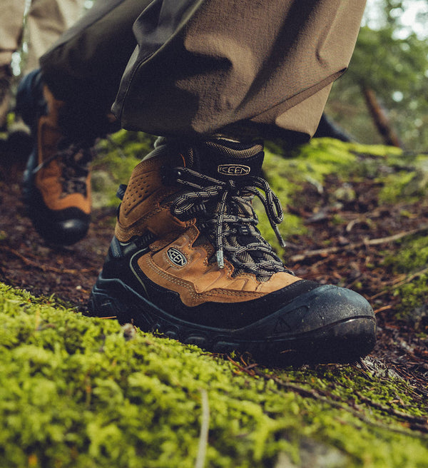 A person wearing a pair of brown and black KEEN wide hiking boots on a forest trail.