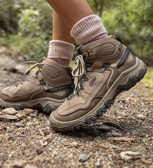 Knee-down shot of woman in tan leather hiking boots and pink crew socks walking across dirt trail. 