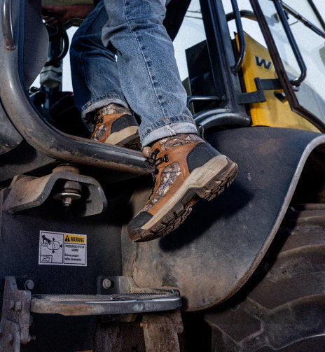 Knee-down shot of man stepping into a large tractor wearing Skokie work boot with camo design detail