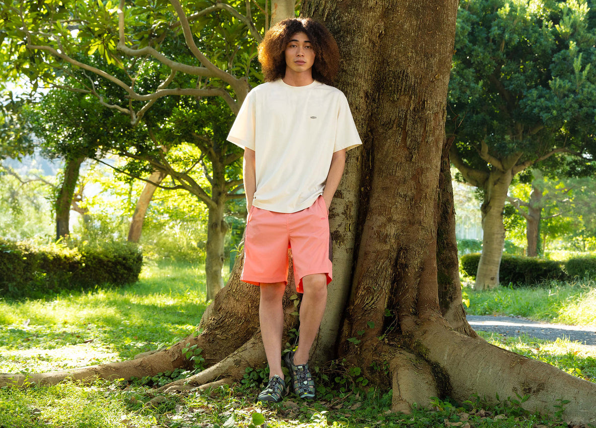 A man in neon pink shorts and bright turquoise Newport sandals standing in front of large tree
