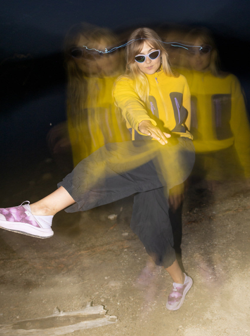 A blurred photo of a woman in yellow jacket, sunglasses, and pink shoes dancing at last light in forest. 
