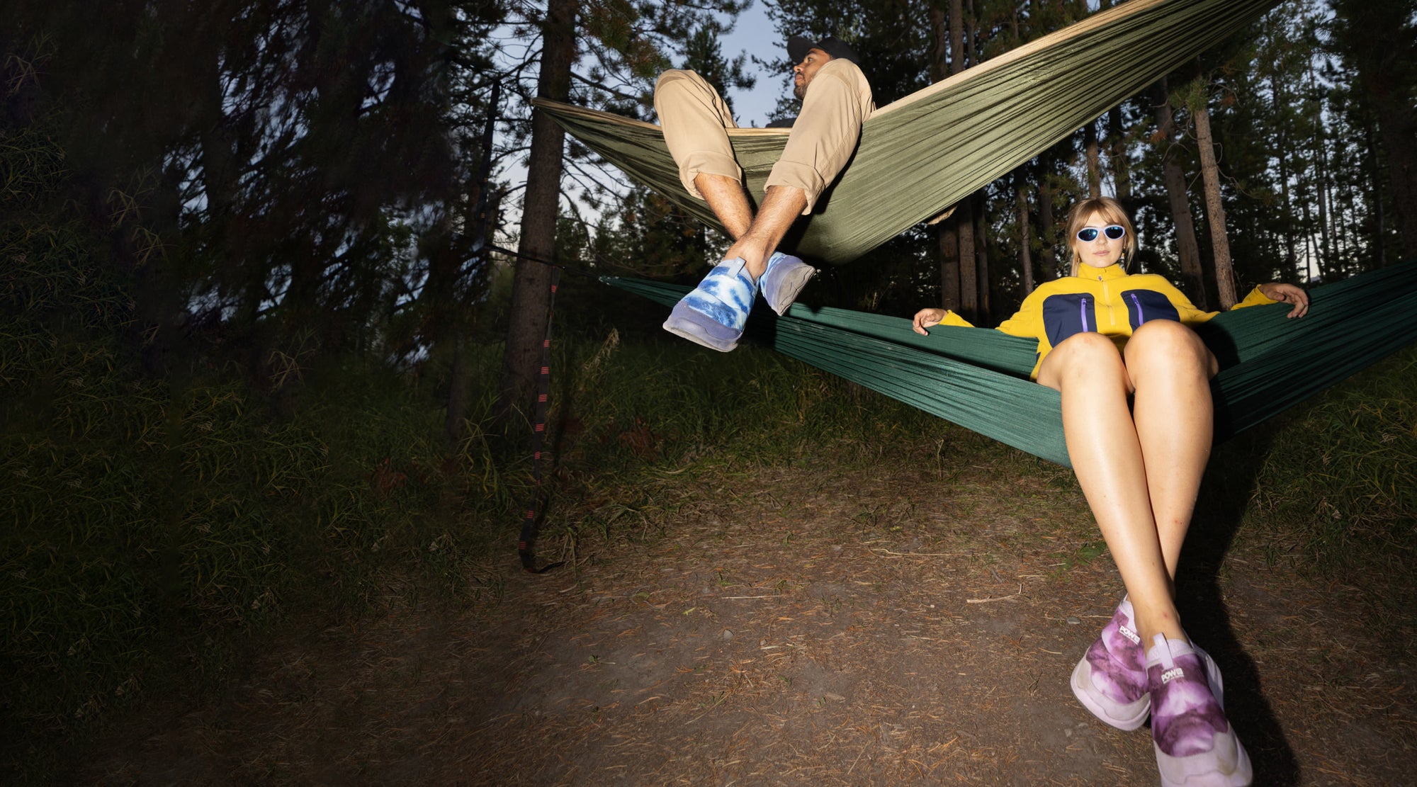 A man and a woman relax together in a hammocks while wearing brightly colored pink and blue  shoes, enjoying a peaceful moment in the forest.