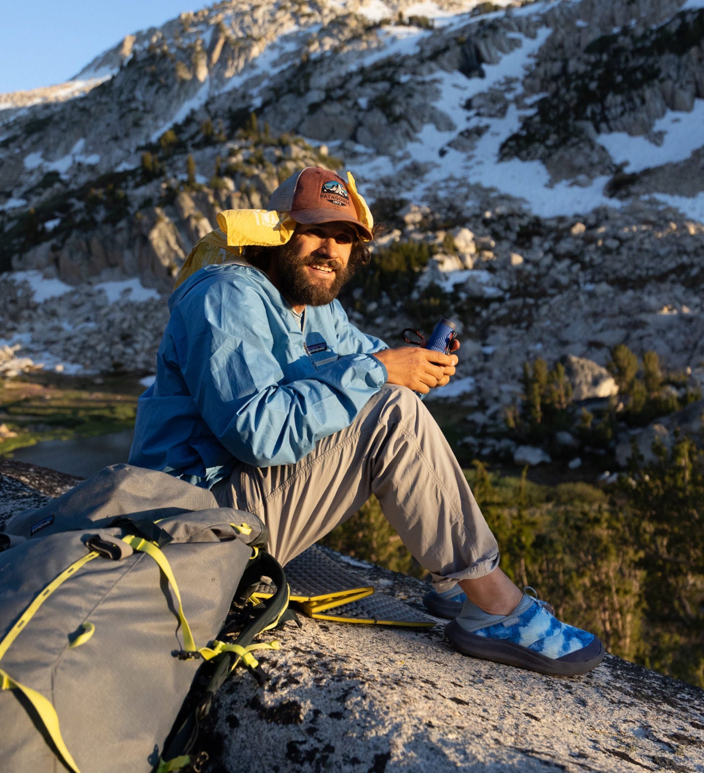 A man in backpacking gear sitting on a rock in front of the mountains, having switched out his hiking boots for cozy blue camp shoes. 